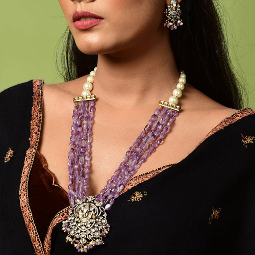 Woman wearing a black saree with gold details and a purple necklace against a green background

